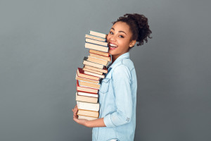 Young african female student isolated on grey wall holding a pile of books standing profile looking camera laughing